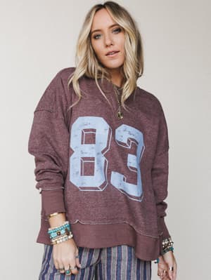 A relaxed, lived-in varsity sweatshirt in washed mocha worn with striped wide-leg pants and layered boho jewelry — the kind of outfit that looks effortless because it actually is.