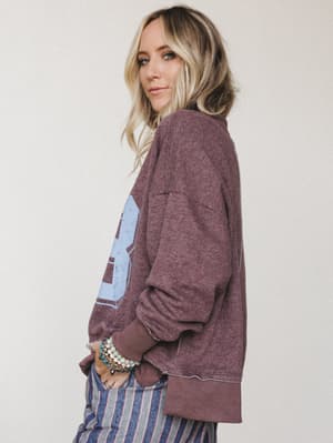A model glances back over her shoulder in an oversized varsity-graphic sweatshirt in a warm plum-mocha, layered over striped wide-leg pants and stacked with beaded and charm bracelets — effortless and a little nostalgic.
