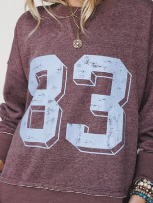 A close-up of a heathered mocha crewneck sweatshirt with a distressed varsity '83' graphic in periwinkle blue, layered with a gold chain necklace and stacked beaded bracelets for that effortlessly collected, lived-in feel.