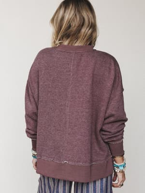 A back-view shot of a relaxed, heathered mauve sweatshirt with visible center seam and raw-edge hem detailing, paired with striped wide-leg pants and layered boho bracelets — the kind of outfit that looks effortless because it actually is.