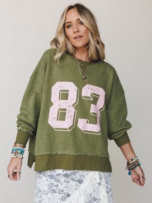 An oversized olive varsity sweatshirt with a distressed pink '83' graphic is styled over a white lace skirt, layered with stacked boho bracelets and a gold chain necklace — that effortless mix of sporty and feminine that just works.