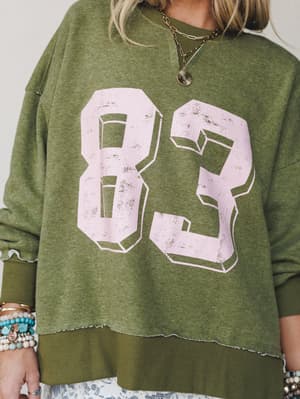 An oversized olive green varsity sweatshirt with a distressed pink '83' graphic, styled with layered gold necklaces, stacked turquoise and beaded bracelets, and a floral hem peeking out beneath — the kind of effortlessly worn-in piece that looks like