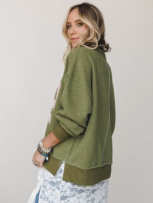 Shot from the back at a three-quarter angle, this oversized olive graphic sweatshirt is styled over a white lace skirt with stacked boho bracelets — the kind of outfit that looks effortless but was absolutely thought about.