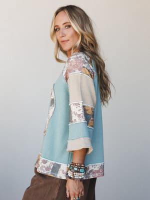 A side-profile shot of a model in a relaxed waffle-knit teal top with patchwork floral panels and a peace sign detail, styled with stacked turquoise and beaded bracelets and brown wide-leg pants — the kind of top that feels like a hug and still looks