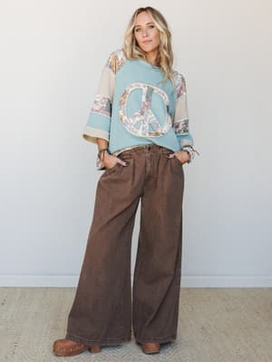 A relaxed, full-body studio shot of a model in a patchwork peace sign thermal top paired with wide-leg chocolate brown pants and wooden clogs — the kind of outfit that feels effortlessly retro without trying too hard.