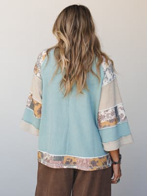 A back-view shot of a relaxed waffle-knit top in soft teal, detailed with patchwork floral print trim along the bell sleeves and hem, paired with wide-leg brown pants — lived-in and effortlessly boho.