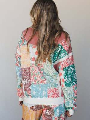 Shot from behind, a model wears a relaxed patchwork sweatshirt bursting with mixed floral, paisley, and botanical prints in a cheerful spring palette — paired with matching wide-leg pants, giving the whole look an easy, collected energy.
