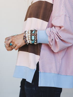 A close-up detail shot of a colorblock and stripe top styled with a stacked bracelet arm party — turquoise stones, pearl beads, and silver rings doing all the right things together.