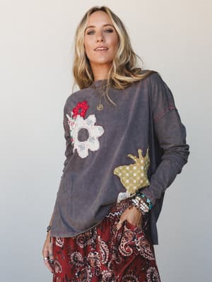 A relaxed, oversized mineral-washed top in ash charcoal gets its personality from mixed-fabric floral and crown appliqués, styled with a burgundy paisley skirt and stacked boho jewelry for a look that feels like it was assembled with intention and a