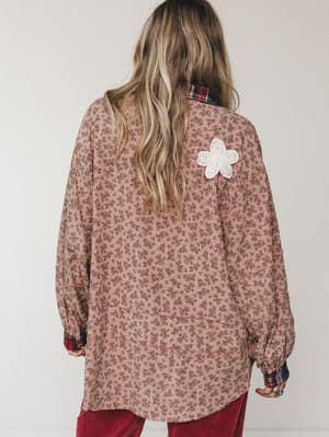 Shot from the back, this oversized floral top in dusty mauve layers over a plaid shirt with a sweet crochet butterfly appliqué on the shoulder — the kind of piece that looks like it's been loved for years.