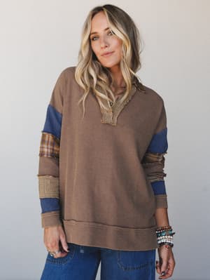 A relaxed waffle-knit pullover top in warm camel with patchwork color-blocked sleeves in cobalt blue, plaid, and gingham — styled with wide-leg denim and stacked beaded bracelets for an effortlessly lived-in fall look.