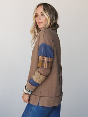 A side-profile shot of a model in a cozy patchwork thermal top with mixed-fabric sleeves — waffle knit, plaid, and denim panels layered together in that perfectly worn-in, collected way — paired with straight-leg jeans and a stack of boho bracelets.