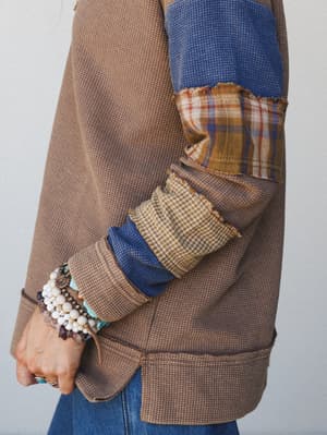 A close-up detail shot of a patchwork thermal top in camel and blue, layered with mixed fabric panels — plaid, gingham, and waffle knit — finished with raw edges and stacked beaded bracelets that feel like they've been collected over years.