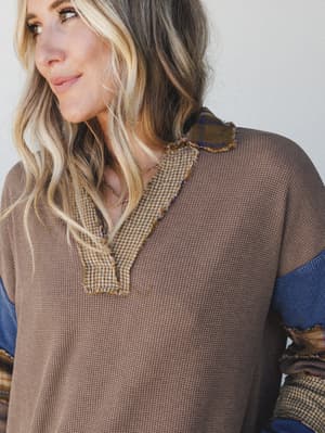A close-up model shot of a relaxed waffle-knit top in warm camel, detailed with mixed-fabric patchwork trim at the v-neckline and contrast colorblock sleeves in denim blue and olive plaid — the kind of top that looks like it has a story.