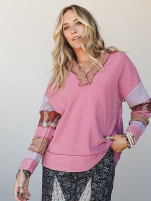 A relaxed, patchwork-style waffle knit top in pink with mixed plaid and gingham contrast panels at the sleeves and collar, styled with dark floral pants and layered boho jewelry — the kind of top that looks like it came together on purpose but feels