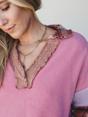 A close-up detail shot of a pink waffle-knit top with a mixed-fabric frayed v-neckline in gingham and plaid, layered gold chain necklaces adding an effortlessly personal touch.