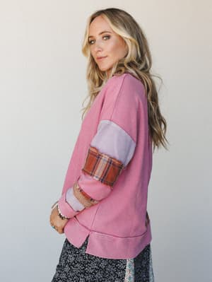 A model shot in profile showing a cozy patchwork pullover top in pink waffle knit with mixed fabric sleeve panels — lavender colorblock, plaid trim, and raw-edge detailing — layered over a floral midi skirt, styled against a soft neutral studio backd