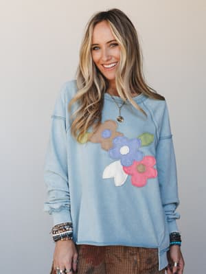 A smiling model wears an oversized dusty blue sweatshirt with bold floral appliqué patches in taupe, periwinkle, pink, and white, styled with stacked beaded bracelets and a delicate gold necklace — casual and expressive with a retro-boho heart.