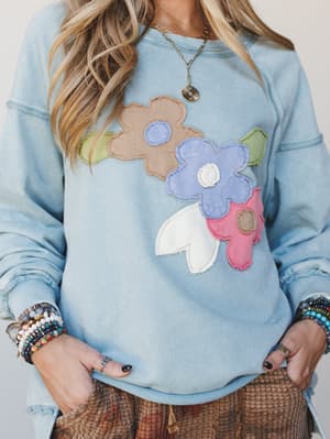 A washed dusty blue sweatshirt with chunky floral appliqué in muted tones of taupe, periwinkle, rose, and cream — styled with stacked beaded bracelets and a gold pendant necklace for that perfectly lived-in, boho-everyday feel.