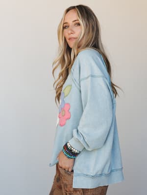 A relaxed, oversized sweatshirt in washed dusty blue with retro floral appliqué — styled with stacked beaded bracelets and printed wide-leg pants for a lived-in, playful everyday look.