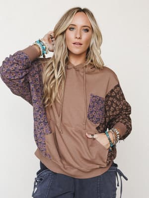 A relaxed, color-blocked hoodie in warm mocha with mixed floral print panels on the sleeves and chest pocket, styled with navy cargo pants and layered boho jewelry for an effortlessly lived-in look.