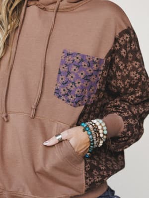 A close-up shot of a mixed-fabric hoodie in warm mocha, featuring floral-printed sleeves and a contrast purple floral chest pocket — styled with a stack of beaded boho bracelets tucked into the kangaroo pocket.