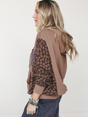 A model photographed from the side showing the back detail of a mixed-media hoodie — warm mocha fleece panels contrast with a dark floral-print body and sleeve, styled with dark joggers and a stack of beaded bracelets for that effortlessly lived-in f