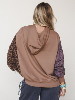 Shot from behind, a mixed-media hoodie with a solid mocha fleece body and contrasting floral-print sleeves in dusty purple and dark brown — styled with navy cargo pants and a stack of turquoise bracelets for that collected, lived-in boho feel.