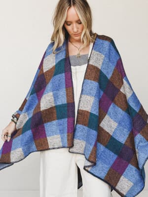A cozy, oversized blanket poncho in a bold color-block plaid drapes wide over white wide-leg pants and a layered top — styled with stacked jewelry and a relaxed, inward gaze that feels like a Saturday morning with nowhere to be.