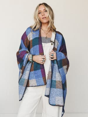 A relaxed, expressive poncho wrap in a bold color-block plaid draped over a white tank and wide-leg pants — layered with turquoise jewelry and silver rings, the whole look feels like a cozy, well-traveled afternoon.