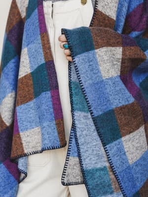 A close-up detail shot of a cozy plaid poncho in rich jewel tones layered over cream wide-leg pants, with a turquoise stone ring peeking through — the kind of outfit that makes a Tuesday feel intentional.