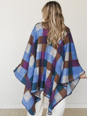 Shot from behind, a model wears a cozy oversized poncho in a bold color-block plaid — periwinkle blue, plum, teal, and warm brown — with whipstitch trim along the hem, paired with white wide-leg pants for a relaxed, lived-in feel.