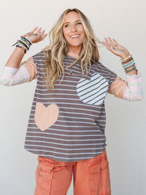 A smiling blonde model poses playfully in a striped mixed-media top with heart appliqués and contrast striped sleeves, paired with terracotta cargo pants and stacked bead bracelets — the whole look has that effortless, personality-first energy.