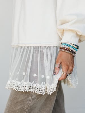 A close-up detail shot of an ivory sweatshirt with a sheer tulle hem extension trimmed in floral lace, styled with corduroy pants and a wrist full of stacked beaded bracelets and silver rings — the kind of layered, personal details that make an outfi