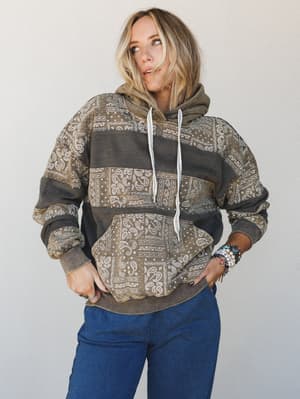 A relaxed, oversized paisley patchwork hoodie in earthy olive and charcoal tones styled with dark blue pants and layered boho bracelets — the kind of sweatshirt that looks like it has a whole personality already.