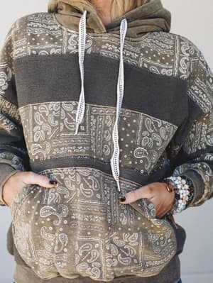 A close-up torso shot of a washed olive and charcoal hoodie sweatshirt in a bandana paisley patchwork print, styled with stacked beaded bracelets and hands tucked into the kangaroo pocket — the kind of cozy you plan your whole Saturday around.