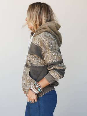 A side-profile shot of a model wearing a cozy patchwork paisley hoodie in washed olive and charcoal tones, styled with stacked turquoise and pearl bracelets — the kind of sweater that looks like it has a whole backstory.