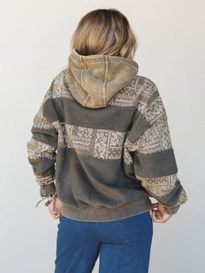 Shot from the back, a model wears an oversized patchwork hoodie in washed olive and charcoal, layered with bandana-print panels across the sleeves and body — styled with blue joggers and stacked bead bracelets for an effortlessly lived-in feel.