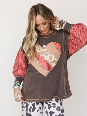 A washed ash brown sweatshirt top with contrast red plaid puff sleeves and a patchwork heart graphic center-front, styled with cow print pants and layered boho jewelry for a playful, lived-in look.