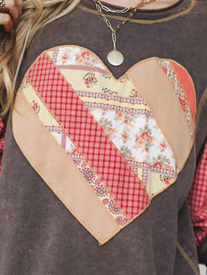 A close-up detail shot of a washed ash sweatshirt with a large patchwork heart graphic made up of mixed floral, gingham, and paisley fabric strips, styled with layered gold chain necklaces and a plaid underlayer peeking through.