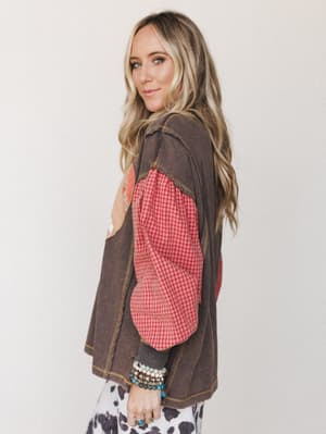 A side-angle model shot showing a layered boho look — a washed charcoal vest with patchwork detailing layered over a red gingham long-sleeve top, finished with stacked turquoise and beaded bracelets and a hint of cow print at the hem.