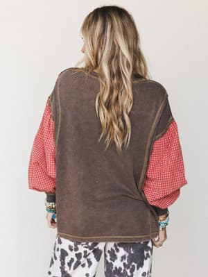 Shot from the back, this layered mixed-media top pairs a washed charcoal sweatshirt-style body with voluminous red gingham sleeves and cow print pants — the kind of outfit that feels effortlessly collected, like she's been dressing this way her whole