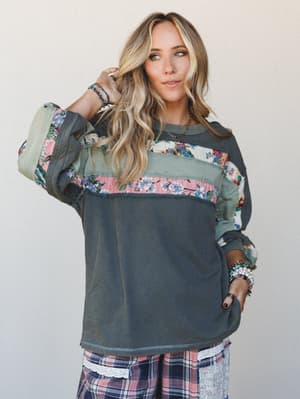 A lived-in, patchwork pullover top in washed charcoal gets a boho upgrade with floral tapestry and sage fabric paneling — layered over plaid wide-leg pants with lace trim, and stacked with bracelets and a pendant necklace for that effortlessly collec