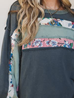 A close-up of a lived-in long sleeve top in washed ash navy, patched with horizontal bands of floral tapestry fabric in blush pink and cream — the kind of piece that looks like it has a history and a personality.