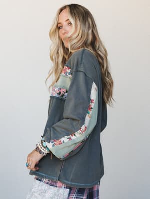 A side-profile shot of a model in a washed ash navy pullover top with patchwork floral and sage fabric panels running down the sleeves — layered over a plaid bottom and accessorized with stacked turquoise and pearl bracelets, giving it that collected