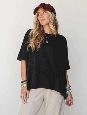 An oversized washed black peplum tee styled with relaxed sand-toned pants, layered beaded bracelets, and a red plaid newsboy cap — the kind of effortless, personality-forward outfit that looks thrown together but really isn't.