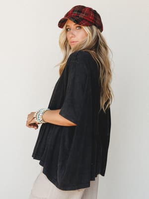 A relaxed oversized tee with a ruffle-hem silhouette shot from the back-angle, styled with a red plaid newsboy cap and stacked bead bracelets — the kind of effortless look that makes a Tuesday feel intentional.