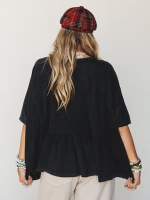 Shot from behind, a model wears a relaxed washed-black tiered ruffle top with a plaid newsboy cap and stacked beaded bracelets — the kind of effortless, lived-in look that feels like it came together without trying.