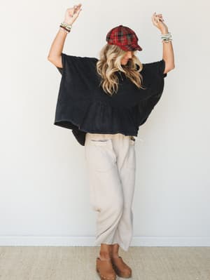 A model with arms raised and a smile-down energy wears an oversized washed black ruffle-hem top paired with cream textured wide-leg pants, a red plaid cap, and tan clogs — the whole look reads like a cozy Saturday that somehow got stylish.