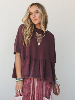 A relaxed, oversized peplum tee in washed mulberry layered over a printed boho skirt — styled with stacked bracelets, a statement coin necklace, and rings, giving the whole look that effortless 'threw it on and somehow looked great' energy.